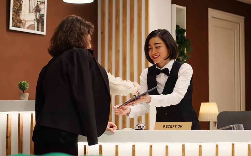 Business travelling guest checking in posh hotel, being helped by bellboy with baggage and receiving room key from receptionist. Resort visitor in professional trip assisted by concierge personnel