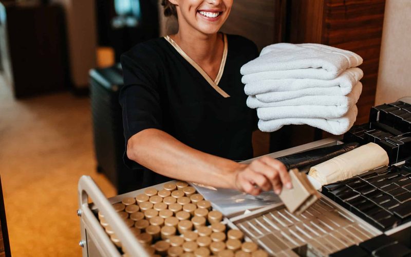 Beautiful young hotel chambermaid in uniform bringing clean towels and other supplies to hotel room.