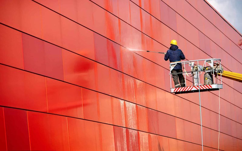 worker of Professional Facade Cleaning Services washing the red wall. Worker wearing safety harness washes wall facade at height on modern building in a crane.